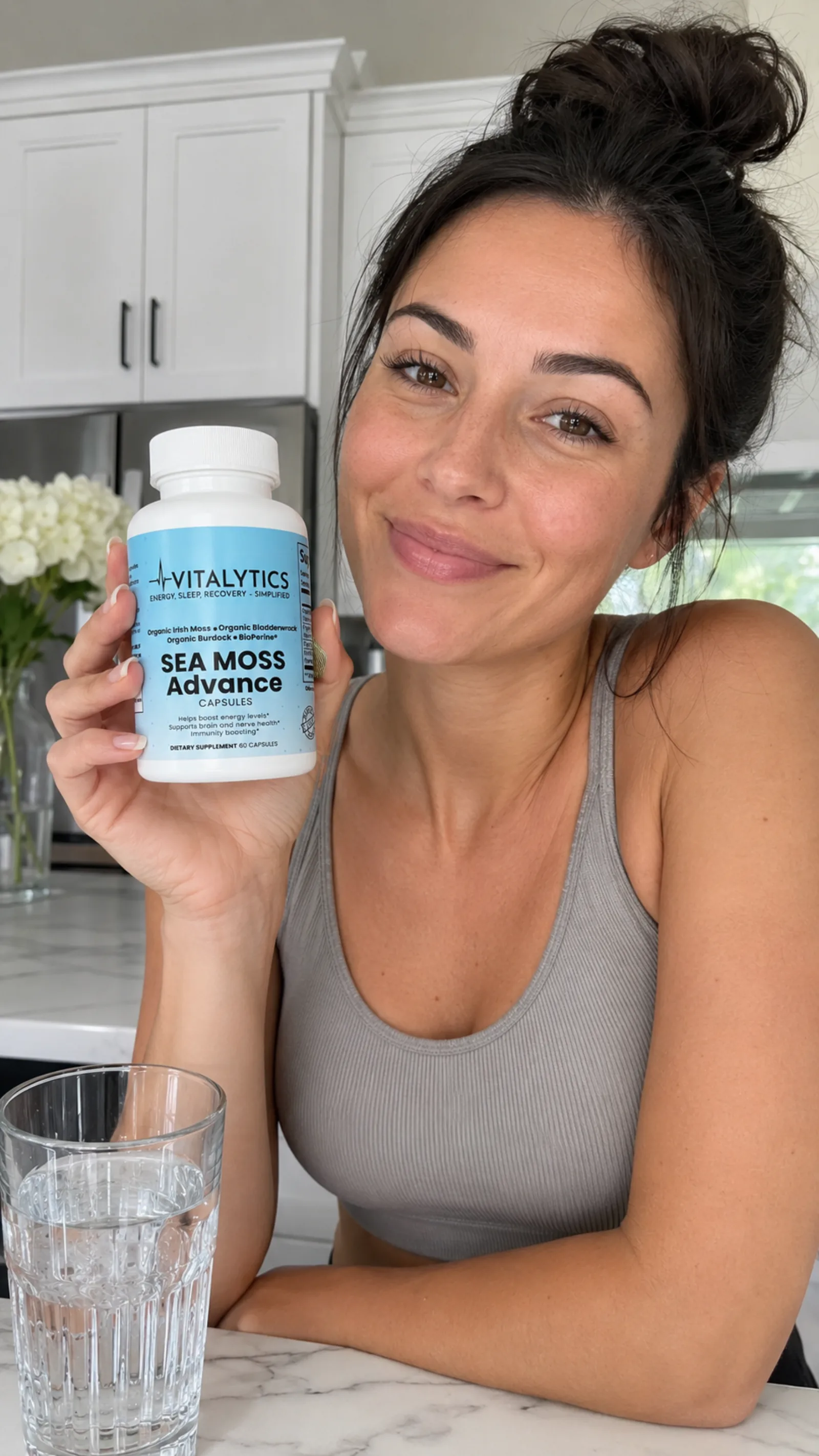 Woman in a bright kitchen holding Vitalytics Sea Moss Advance next to a glass of water.