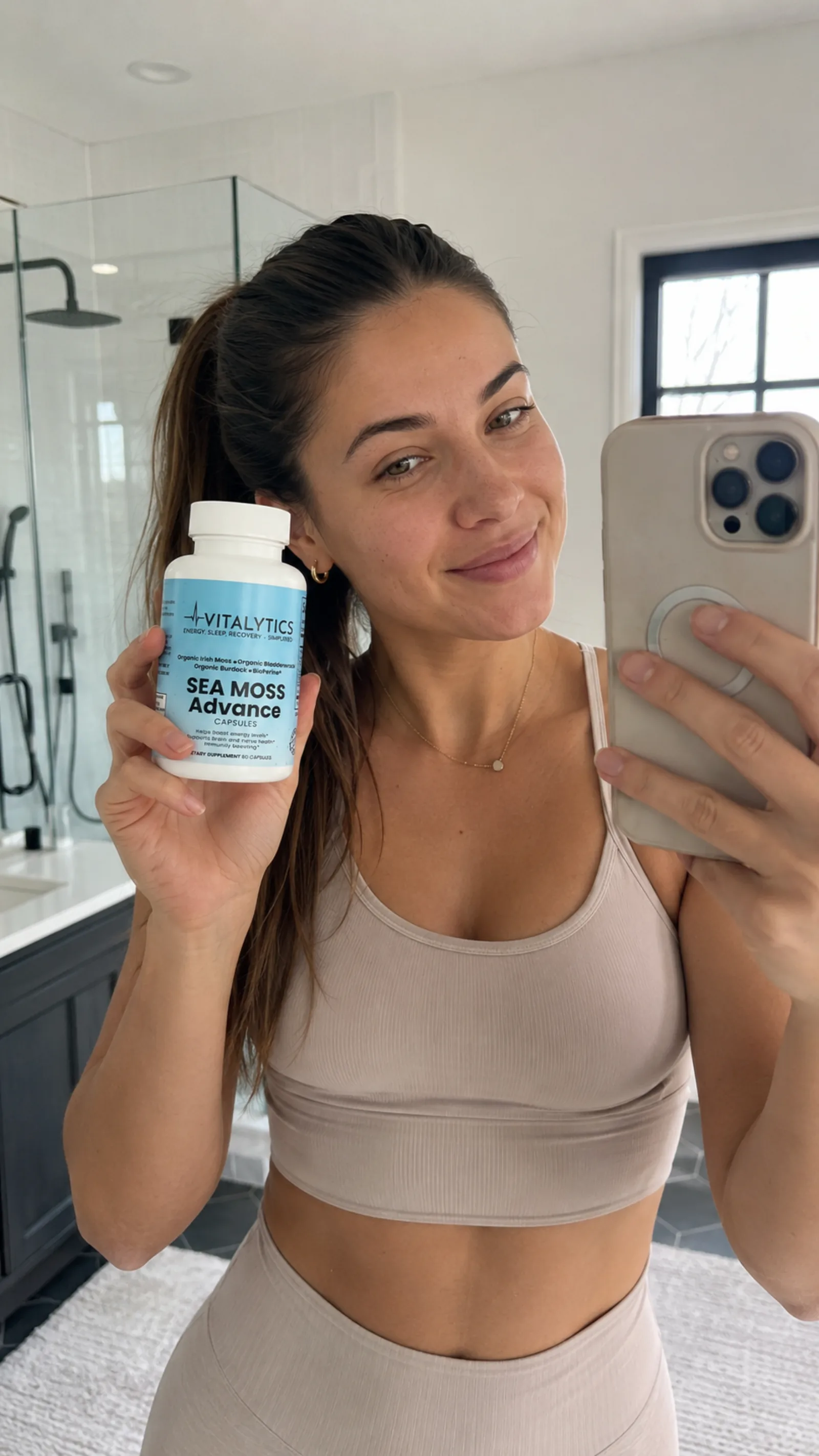 Woman taking a mirror selfie while holding Vitalytics Sea Moss Advance in a bright bathroom.